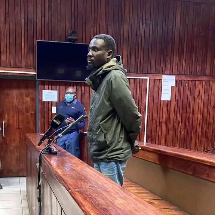 A serious-looking man in an olive jacket stands with hands behind his back in a wood-paneled courtroom, near microphones and a masked official.