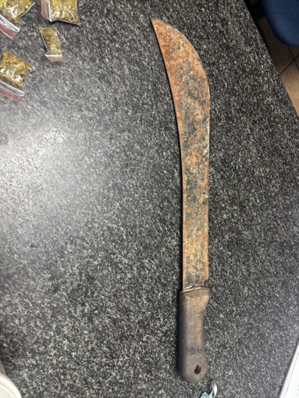 A heavily rusted machete with a dark handle lies on a speckled gray surface beside small packets, creating a worn, utilitarian impression.
