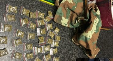 An evidence display shows dozens of small sealed packets, camouflage clothing, labeled bags, and a curved blade on a dark floor, suggesting a police seizure.