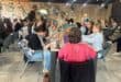 People paint together at a long table in a warmly lit art studio, chatting as brushes and colorful canvases create a relaxed, social atmosphere.