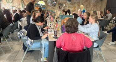 People paint together at a long table in a warmly lit art studio, chatting as brushes and colorful canvases create a relaxed, social atmosphere.
