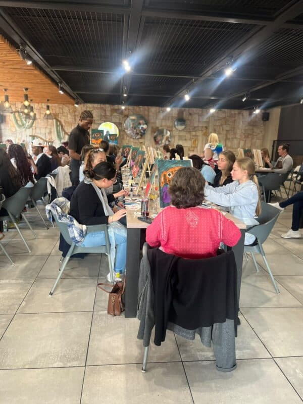 People paint together at a long table in a warmly lit art studio, chatting as brushes and colorful canvases create a relaxed, social atmosphere.