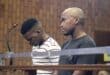Two men stand behind a courtroom barrier: one bows his head beside a microphone while the other faces forward, both appearing solemn against wood paneling.