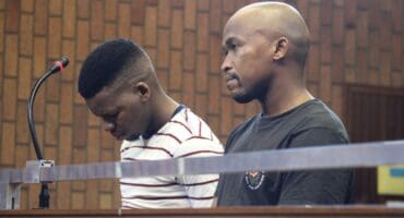 Two men stand behind a courtroom barrier: one bows his head beside a microphone while the other faces forward, both appearing solemn against wood paneling.