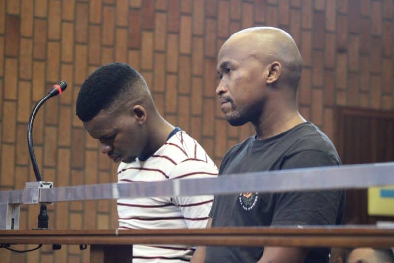 Two men stand behind a courtroom barrier: one bows his head beside a microphone while the other faces forward, both appearing solemn against wood paneling.