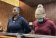 Two women stand behind a courtroom-style wooden rail; one faces forward while the other, wearing a knit hat and mask, looks down. The mood is solemn.