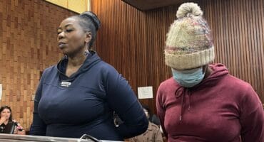 Two women stand behind a courtroom-style wooden rail; one faces forward while the other, wearing a knit hat and mask, looks down. The mood is solemn.