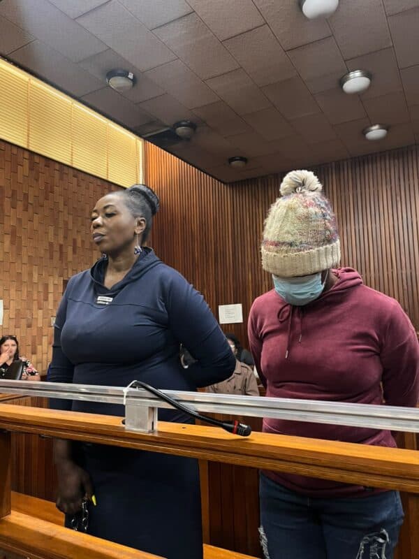 Two women stand behind a courtroom-style wooden rail; one faces forward while the other, wearing a knit hat and mask, looks down. The mood is solemn.
