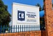 A sunny view of the Con Amore School sign behind a red-brick wall and blue iron fence, beneath a clear blue sky; the scene feels welcoming.