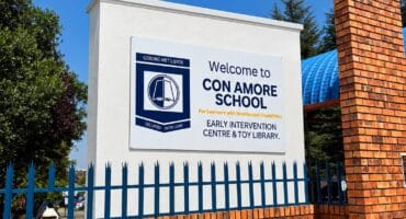 A sunny view of the Con Amore School sign behind a red-brick wall and blue iron fence, beneath a clear blue sky; the scene feels welcoming.