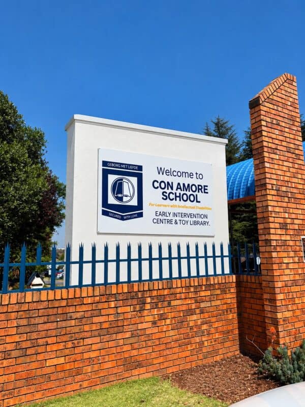 A sunny view of the Con Amore School sign behind a red-brick wall and blue iron fence, beneath a clear blue sky; the scene feels welcoming.