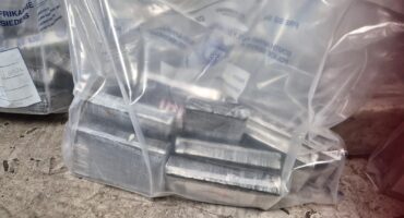 Clear labeled plastic bags contain stacked rectangular metal components on a worn concrete floor, creating an industrial, utilitarian scene.