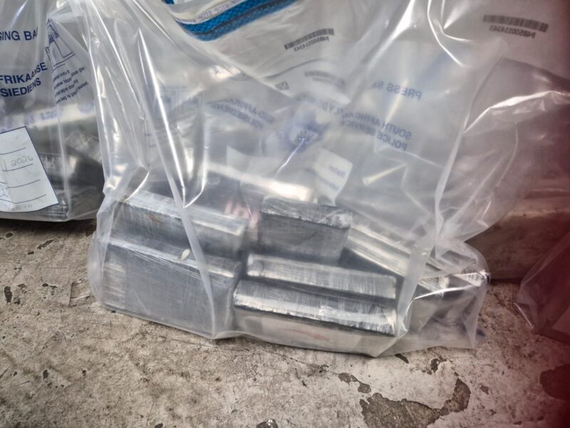 Clear labeled plastic bags contain stacked rectangular metal components on a worn concrete floor, creating an industrial, utilitarian scene.