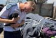 A uniformed police officer gently cradles a small black dog beside blankets and belongings at an outdoor shelter, conveying compassion amid hardship.