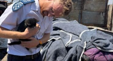 A uniformed police officer gently cradles a small black dog beside blankets and belongings at an outdoor shelter, conveying compassion amid hardship.