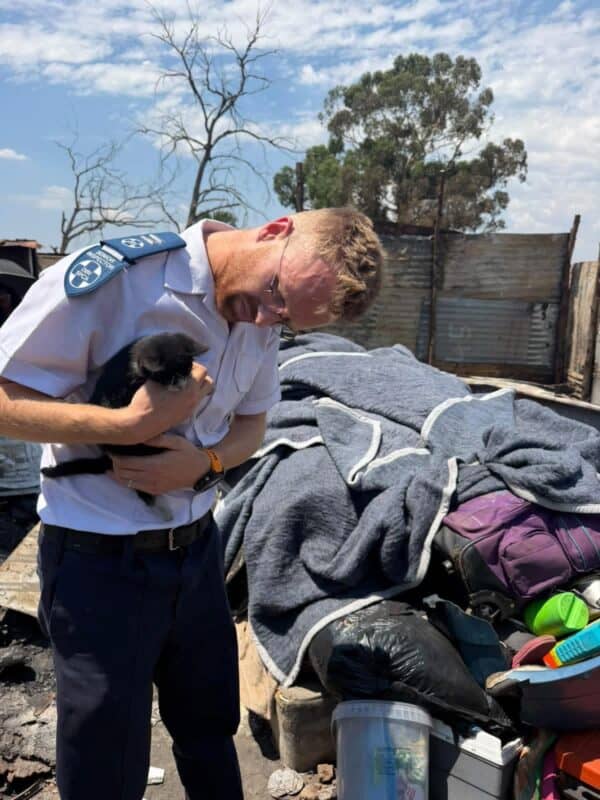 A uniformed police officer gently cradles a small black dog beside blankets and belongings at an outdoor shelter, conveying compassion amid hardship.