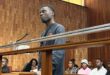 A young man stands at the microphone in a wood-paneled courtroom, with seated people behind him; the scene feels formal and tense.
