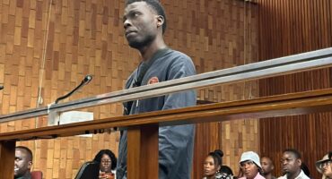 A young man stands at the microphone in a wood-paneled courtroom, with seated people behind him; the scene feels formal and tense.