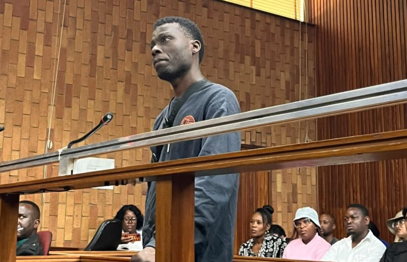 A young man stands at the microphone in a wood-paneled courtroom, with seated people behind him; the scene feels formal and tense.