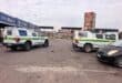 Two blue-and-yellow police vehicles are parked in a paved lot beside a market, with a civilian car in the foreground; the scene feels calm and watchful.
