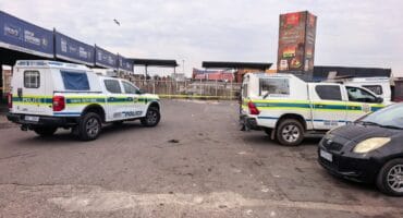 Two blue-and-yellow police vehicles are parked in a paved lot beside a market, with a civilian car in the foreground; the scene feels calm and watchful.