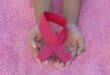 Two cupped hands hold a bright pink awareness ribbon against a textured pink background, symbolizing compassion and support for breast cancer awareness.