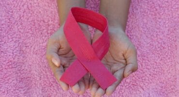 Two cupped hands hold a bright pink awareness ribbon against a textured pink background, symbolizing compassion and support for breast cancer awareness.