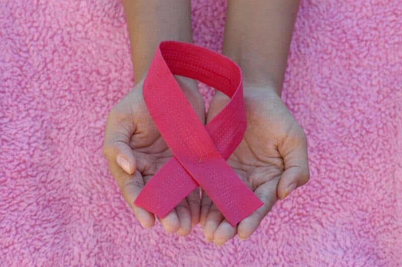 Two cupped hands hold a bright pink awareness ribbon against a textured pink background, symbolizing compassion and support for breast cancer awareness.