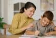 A mother helping her young son with homework at a table, smiling as he writes in a notebook.