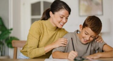 A mother helping her young son with homework at a table, smiling as he writes in a notebook.