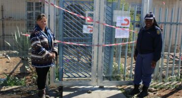 Two women stand in front of a fence with a sign.