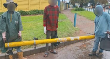 People standing behind a yellow barrier on a rainy sidewalk in Alberton, South Africa.