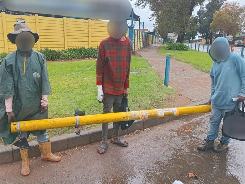 People standing behind a yellow barrier on a rainy sidewalk in Alberton, South Africa.