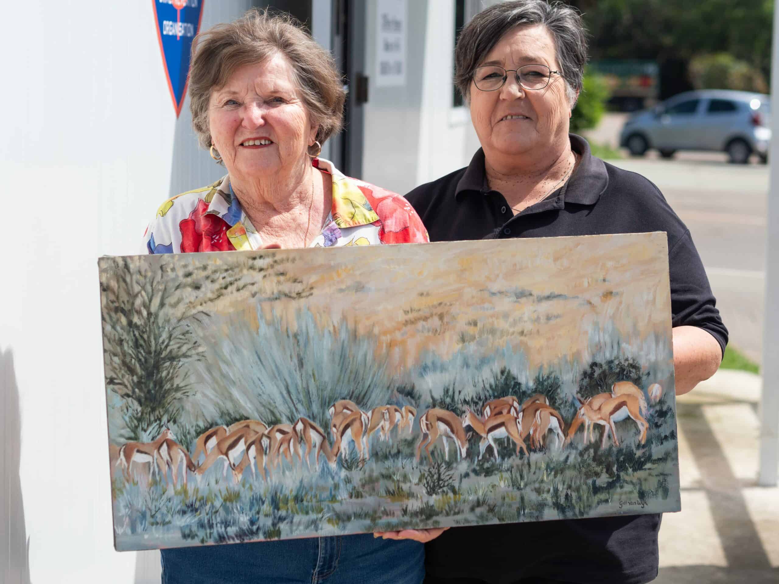 Two woman hold up a painting of Springbok grazing.
