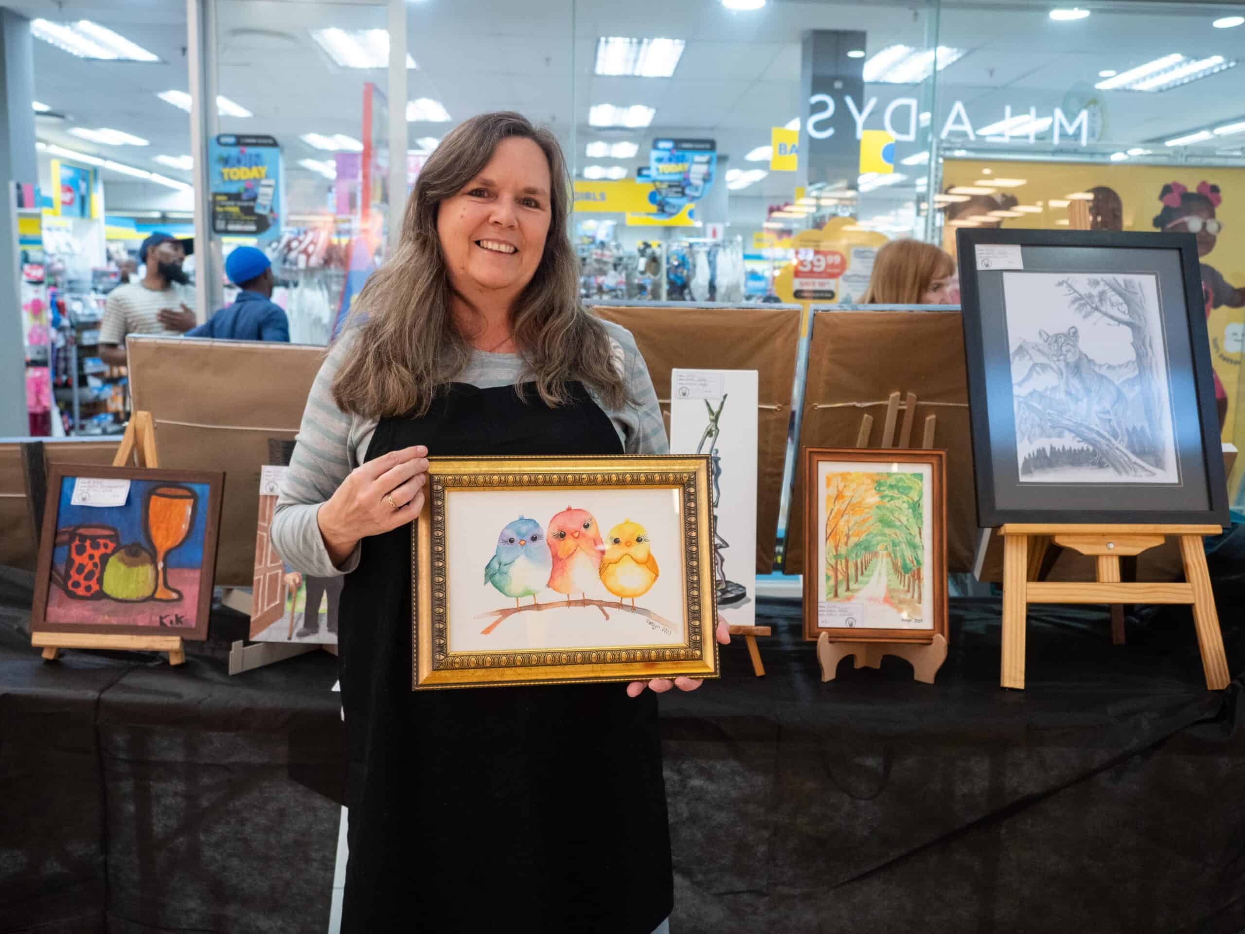 A woman holding her artwork. Behind her is more artwork.