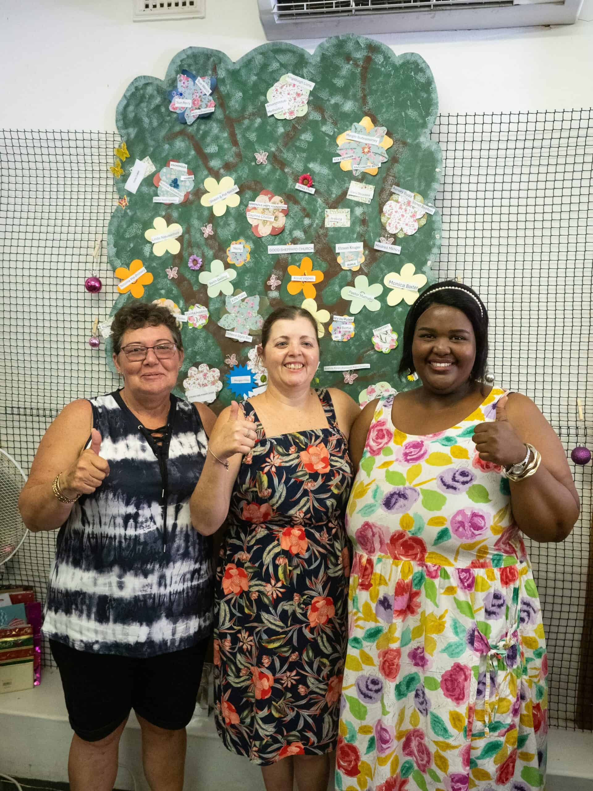 Three woman in front of a donation tree.