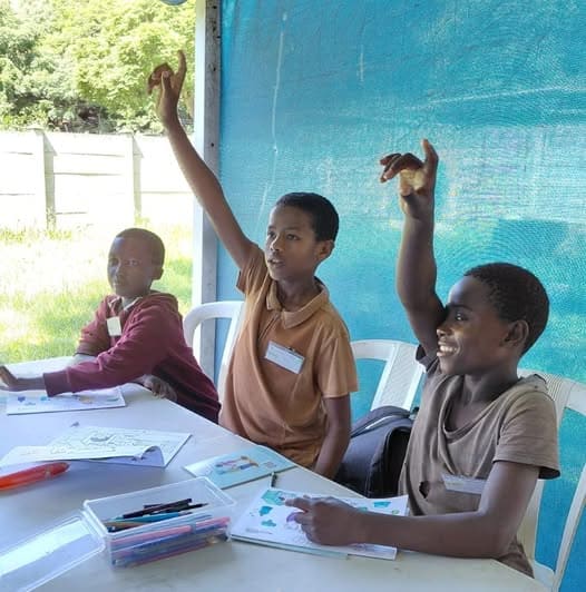 children at an outdoor class, two raising their hands to ask questions
