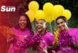 Three women wearing pink t-shirts cheering with pom poms and bright yellow balloons in the background