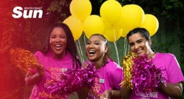 Three women wearing pink t-shirts cheering with pom poms and bright yellow balloons in the background