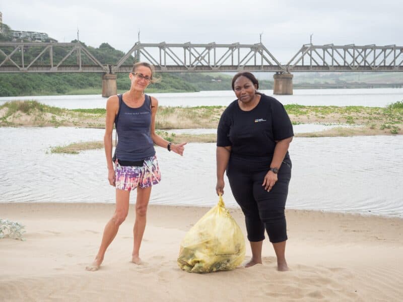 two woman with a bag of litter on the beach, a bridge in the background