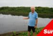 a man standing on a river bank gesturing towards the filth inside the water