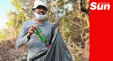 a man holding a black rubbish bag, putting in a glass bottle along a beach promenade