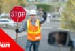 a road traffic officer holding a stop sign. in the foreground is a man visible through his car rearview mirror