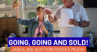 a woman holds up an item up for auction and an auctioneer points to a bidder