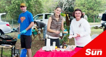 a photograph of three people manning a car boot market stand, two making a hot dog, one working a grill