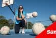 a woman crouches down at a netball field with netballs bouncing all around her