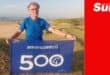 a man on the beach holding up a celebratory banner for his 500th parkrun