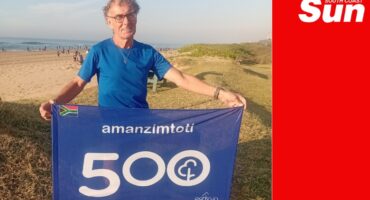 a man on the beach holding up a celebratory banner for his 500th parkrun