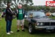 two men shaking hands on a field in front of a classic muscle car