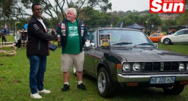 two men shaking hands on a field in front of a classic muscle car